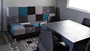 A room with a wooden table, a patterned sofa, a wall clock, and a window. - Mobile Home “Home” with Private Terrace (Plounévézel)