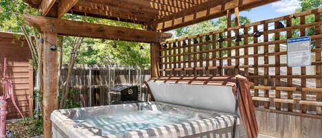 A hot tub under a wooden pergola with a safety sign, surrounded by a wooden fence and greenery.
