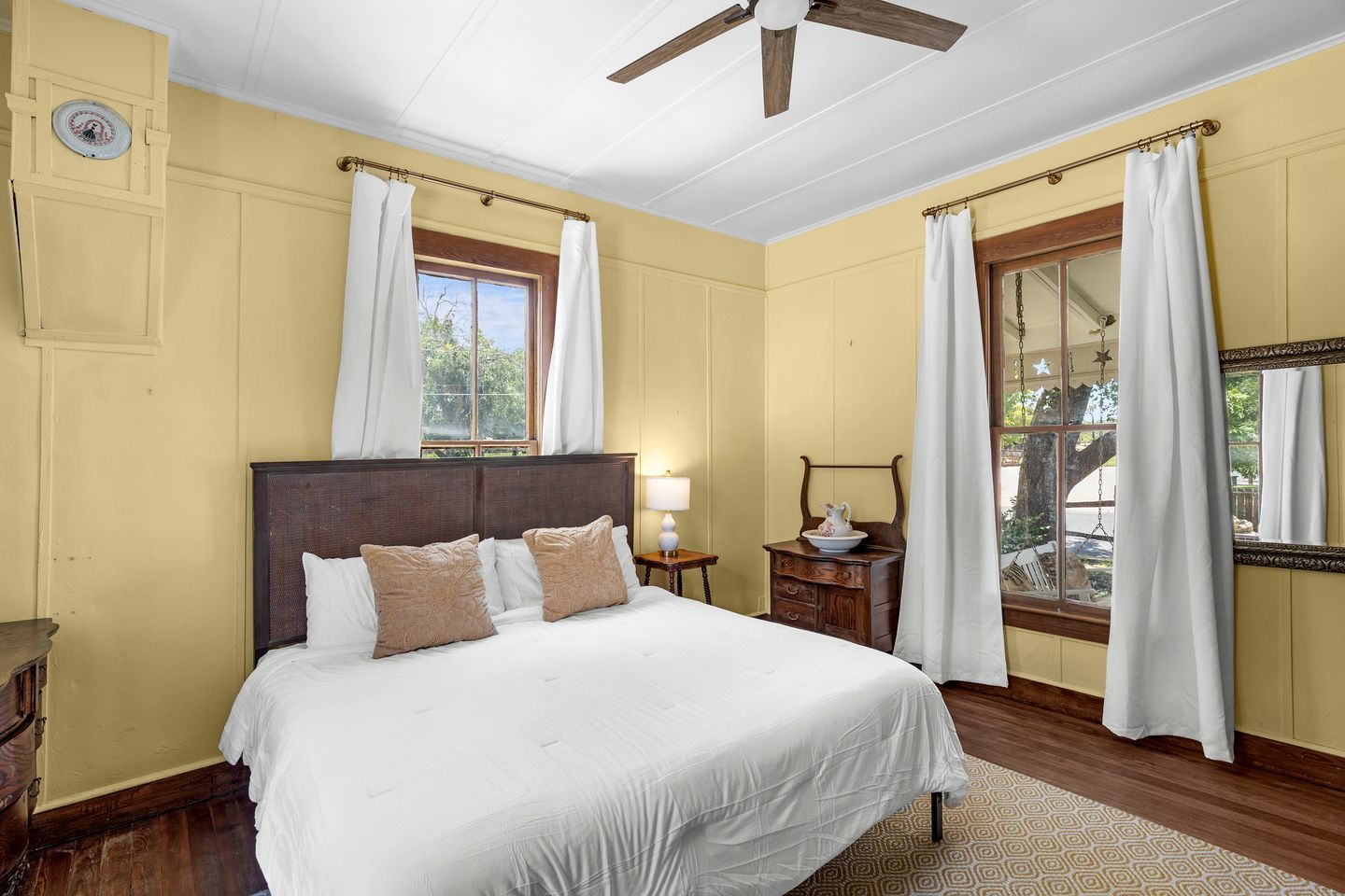 A bedroom with a large bed, a ceiling fan, a window with curtains, and a nightstand with a lamp.