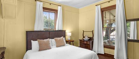 A bedroom with a large bed, a ceiling fan, a window with curtains, and a nightstand with a lamp.