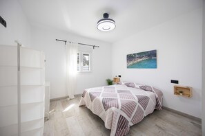 Ein Schlafzimmer mit Bett, einem fensterbelaubten Fenster, einer Deckenleuchte, einer an der Wand befestigten Ablage mit einer Pflanze und einem Bild an der Wand.