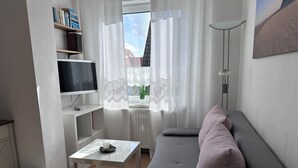 En moderne stue med en grå sofa, et lille hvidt bord, et fjernsyn og en reol.