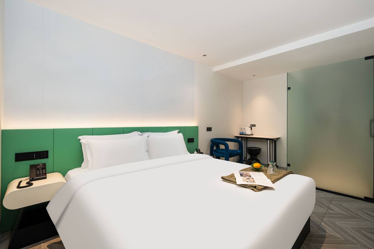 A hotel room with a bed, a desk with a chair, a phone, and a small table with a fruit bowl and a glass.
