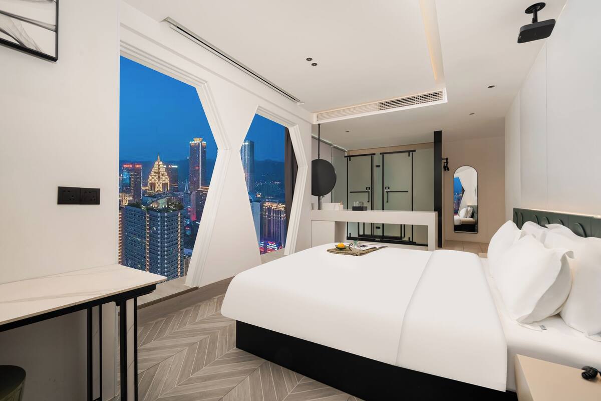 A modern hotel room with a large bed, a desk, and a view of a cityscape at night.