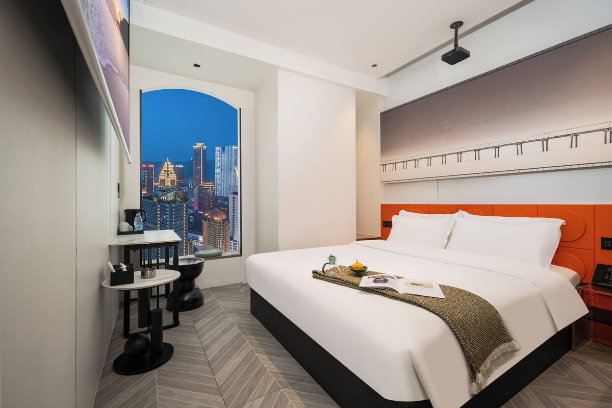A modern hotel room with a large bed, a desk, a chair, and a view of a cityscape.