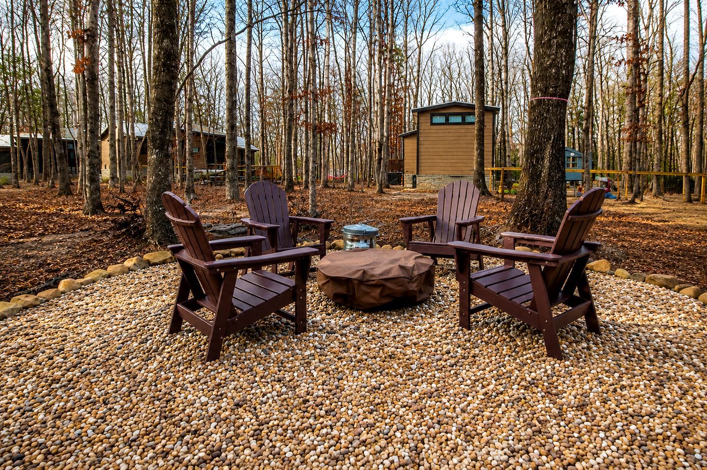 Un espace aménagé avec des fauteuils Adirondack et un foyer, le tout entouré d’une zone boisée.