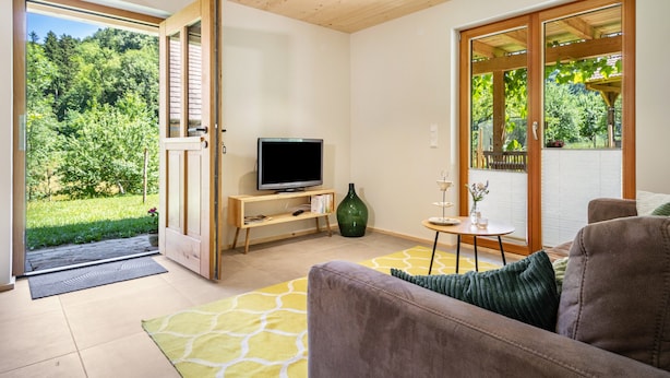 Ein modernes Wohnzimmer mit Fernseher, einer Couch, einem kleinen Tisch und Blick auf einen Garten durch große Fenster.