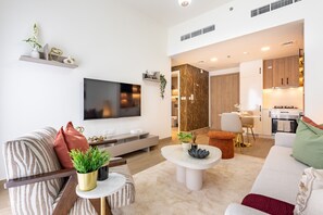 A modern living room with a flat-screen TV, sofa, coffee table, and a kitchen area visible in the background. - Stylish 1BR Just Steps from Town Square Park, Minutes Drive to Miracle Garden (Dubai)