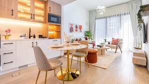 Eine moderne Küche mit weißen Schränken, ein Essbereich mit rundem Tisch und Stühlen sowie ein Wohnzimmer mit Sofa und Fernseher.