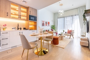 A modern kitchen with white cabinets, a dining area with a round table and chairs, and a living room with a sofa and a TV. - Stylish 1BR Just Steps from Town Square Park, Minutes Drive to Miracle Garden (Dubai)