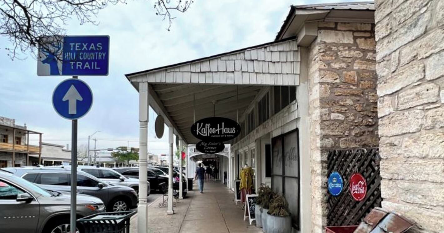A street view with a Texas Hill Country Trail sign, a Kaffe Haus restaurant, parked cars, and a stone building facade.