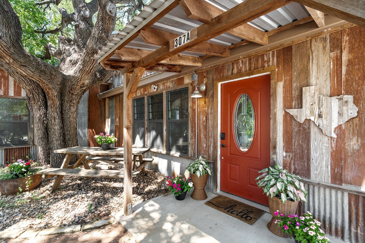 Un bâtiment rustique en bois, avec une porte rouge et le numéro 307A, doté d’un porche couvert avec une table et des chaises, entouré de plantes en pot et d’un grand arbre.