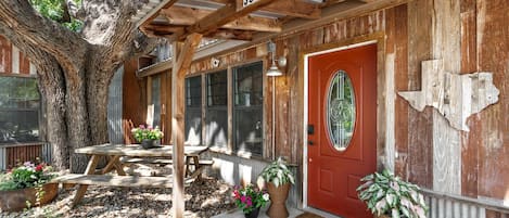 A rustic wooden building with a red door, numbered 307A, featuring a covered porch with a table and chairs, surrounded by potted plants and a large tree.