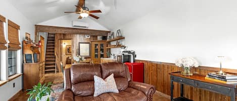 Ein geräumiges Wohnzimmer mit einer Ledersofa, einem hölzernen Deckenventilator und einem Küchenbereich im Hintergrund.
