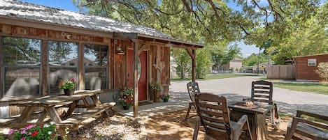 A rustic wooden building with a red door, a covered patio area with wooden furniture, and potted plants.