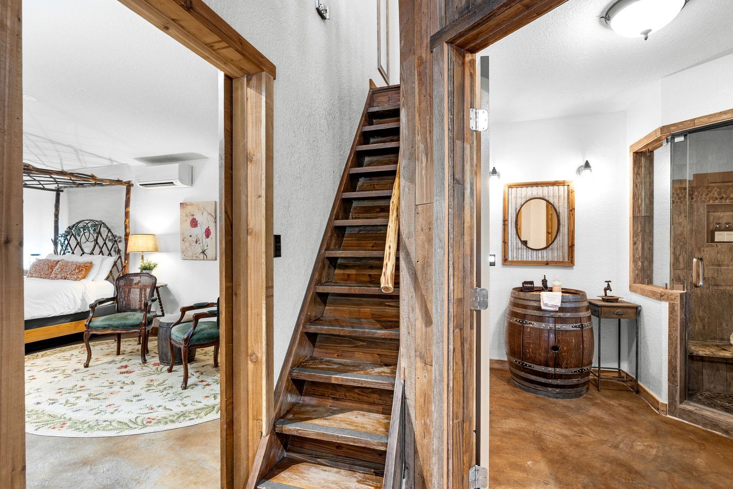 A bedroom with a four-poster bed, a sitting area with chairs, a wooden staircase, a bathroom with a wooden barrel sink, and a large mirror.