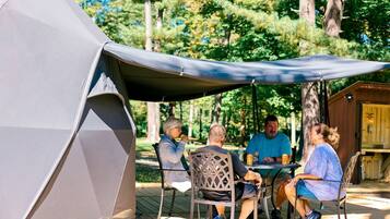 Eine Gruppe von Menschen sitzt an einem Tisch im Freien unter einem Zelt, mit einer Holzhütte im Hintergrund.