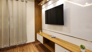 A modern living room with a flat-screen TV mounted on a wall, wooden flooring, and a built-in wooden console table with drawers. - Neelambari Estate Stay (Madikeri)