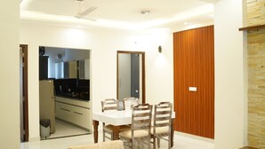 A dining area with a white table and chairs, a kitchen in the background, and a wooden panel wall. - Harmony Haven: Luxurious 3BHK Haven for All. (Sahibzada Ajit Singh Nagar)