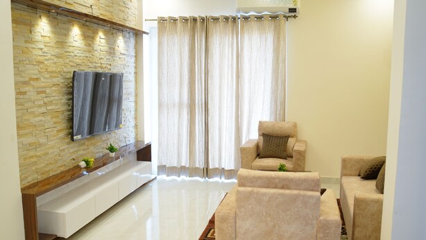A modern living room with a stone wall, a flat-screen TV, beige sofas, and a large window with curtains. - Harmony Haven: Luxurious 3BHK Haven for All. (Sahibzada Ajit Singh Nagar)