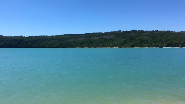 A clear view of a beach with calm water, a distant forested hill, and a clear sky. - Holiday Home "Gîte Des 4 Lacs" with Mountain View, Private Terrace and Wi-Fi (Vertamboz)