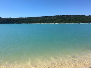 A clear view of a beach with calm water, a distant forested hill, and a clear sky.