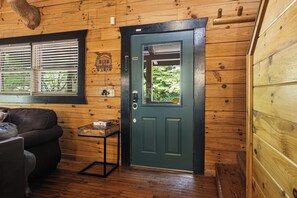 A wooden cabin interior with a green door, a window with blinds, a small table with a box, and a couch. - New Listing!  Hot Tub, Game Room, Theater, Close to Town + Free Tickets (Gatlinburg)