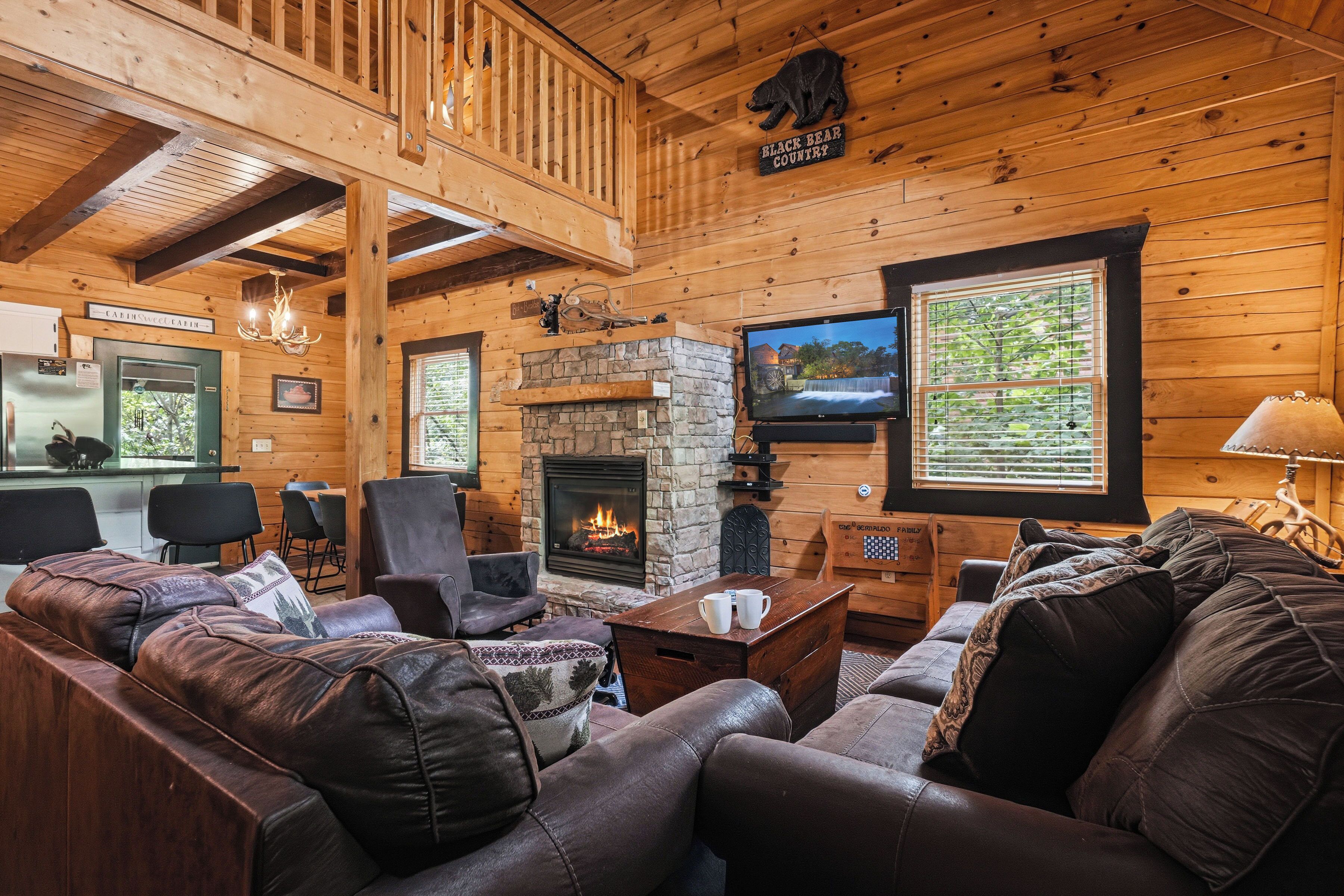 A cozy cabin interior with a stone fireplace, leather sofas, a dining area, and a television mounted on the wall.