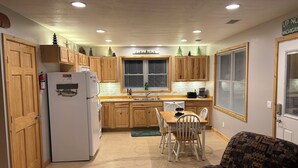 A compact kitchen with wooden cabinets, a white refrigerator, a dining table with chairs, and a patterned armchair. - Amazing hilltop views of gorgeous Sage Lake with beach and boat launch access. (Hale)