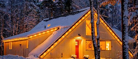 A cabin with a sloped roof, surrounded by snow-covered trees, and illuminated by string lights.