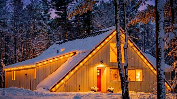 A cabin with a sloped roof, surrounded by snow-covered trees, and illuminated by string lights.
