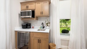 A compact kitchen with wooden cabinets, a stainless steel microwave, and a window with curtains. - Wild Rose Suite (Rome)