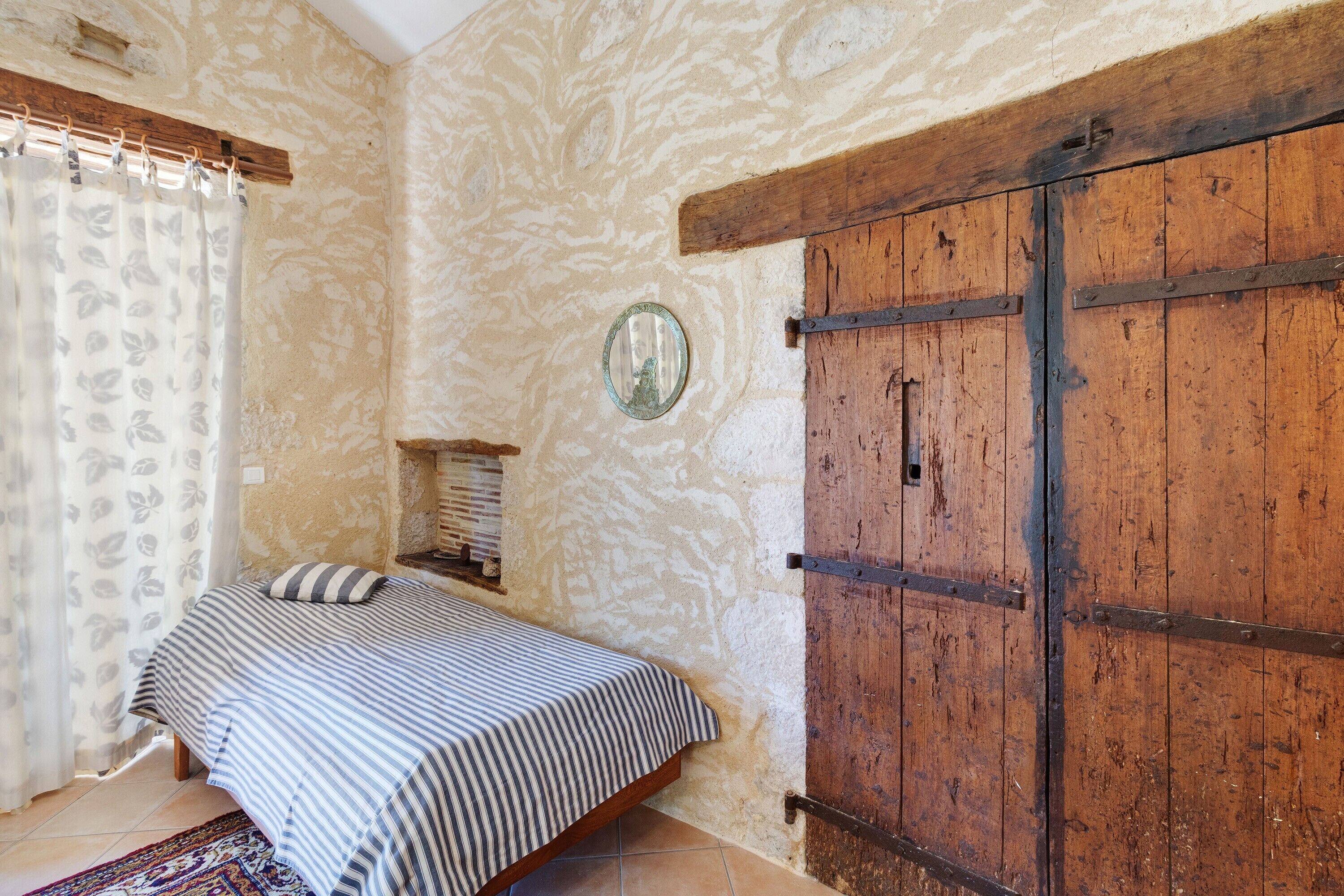 Ein Schlafzimmer mit einem Bett, einem Holzschrank, einem Spiegel und einem Fenster mit Vorhang.