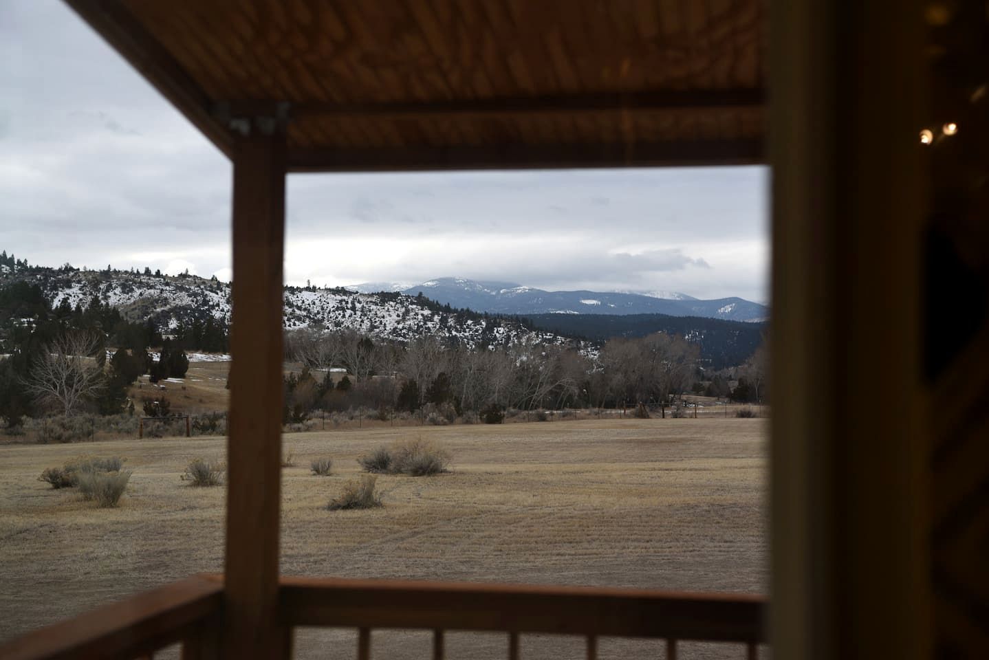 A view from a wooden-framed window overlooking a snowy landscape with trees and mountains.