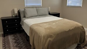 Ein Schlafzimmer mit einem Bett, zwei Nachttischen, einer Kommode und zwei Fenstern mit Jalousien.