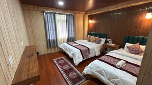 A room with two beds, a wooden floor, a window with curtains, and a nightstand with a lamp.
