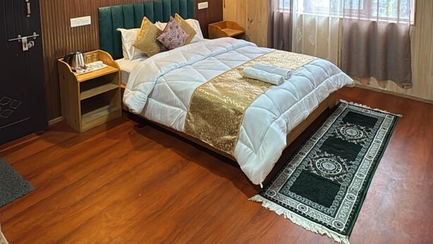 A bedroom with a wooden ceiling, a bed with a white and gold bedspread, two bedside tables, a nightstand, a window with curtains, and a wooden floor.