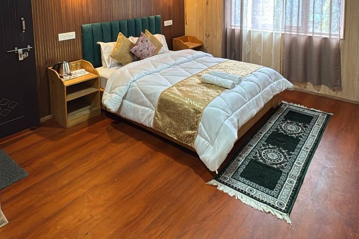A bedroom with a wooden ceiling, a bed with a white and gold bedspread, two bedside tables, a nightstand, a window with curtains, and a wooden floor.