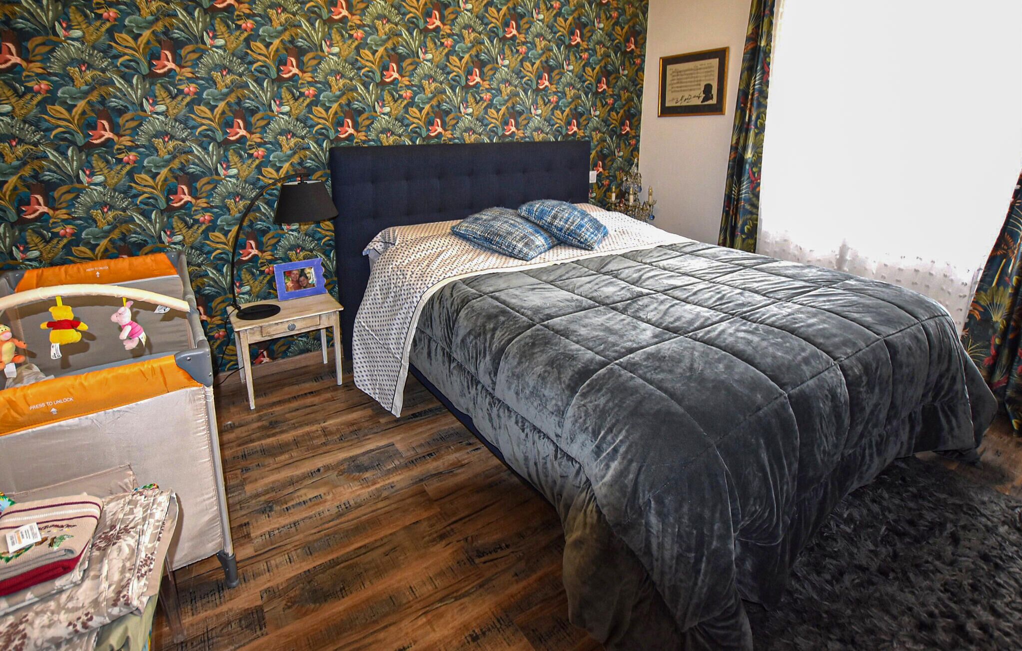 A bedroom with a bed, a nightstand, a small table with toys, a patterned wallpaper, and a framed certificate on the wall.