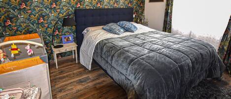 A bedroom with a bed, a nightstand, a small table with toys, a patterned wallpaper, and a framed certificate on the wall.