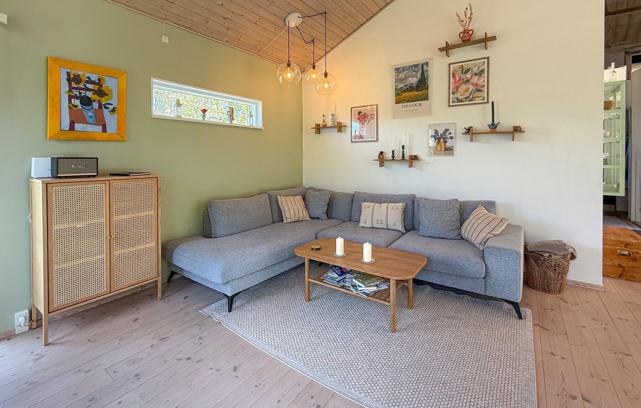 Ein Wohnzimmer mit grauem Sofa, einem Holztisch, einem Korbuscheinmöbel und Wandregalen mit Kunstwerken.