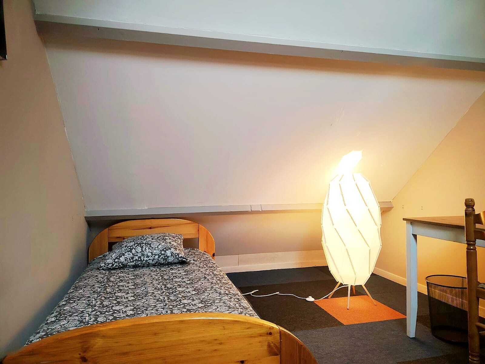 Ein kleines Zimmer mit einem Einzelbett, schrägverdeckter Decke, Nachttisch, Stuhl und Stehlampe.