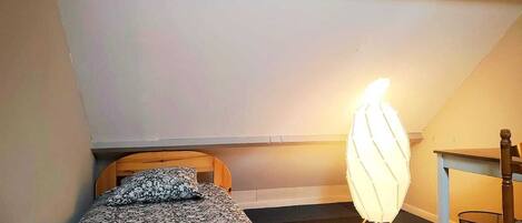 Ein kleines Zimmer mit einem Einzelbett, schrägverdeckter Decke, Nachttisch, Stuhl und Stehlampe.