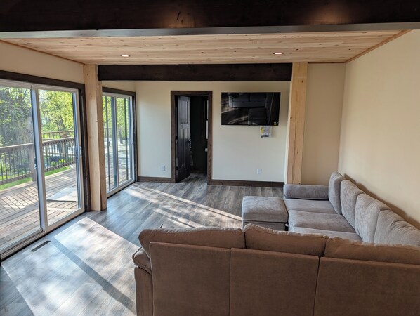 A spacious living area with a large sliding glass door leading to a deck, a grey sofa, and a flat-screen TV mounted on the wall. - Newly Remodeled Lakefront Cabin w/ Hot Tub & Private Dock (Merrifield)