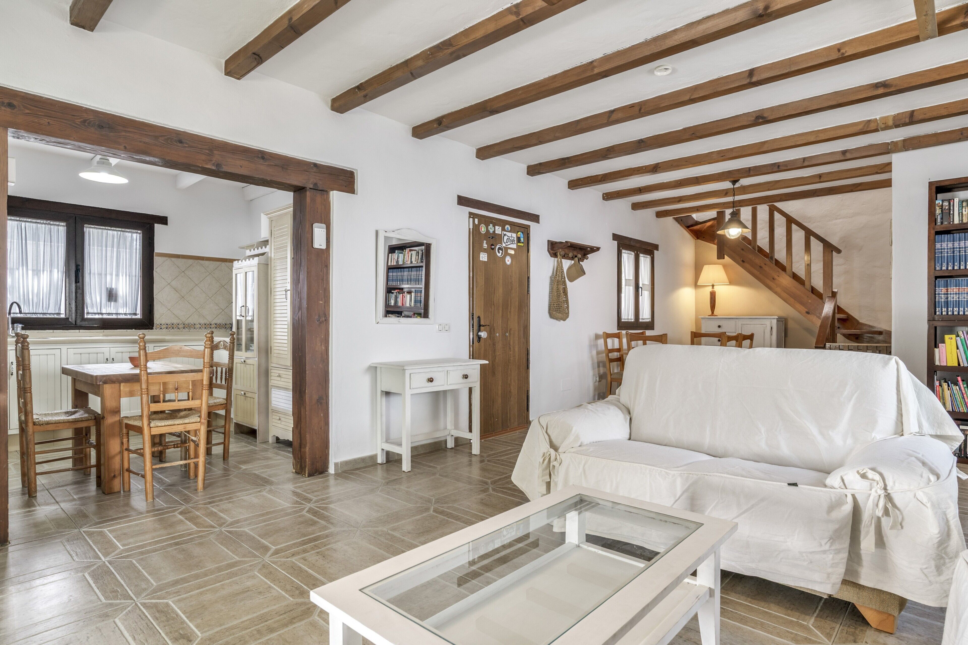 Uma sala de estar acolhedora com um sofá, uma mesa de centro de vidro, vigas de madeira no teto, uma escada e uma área de cozinha com uma mesa de jantar e cadeiras.