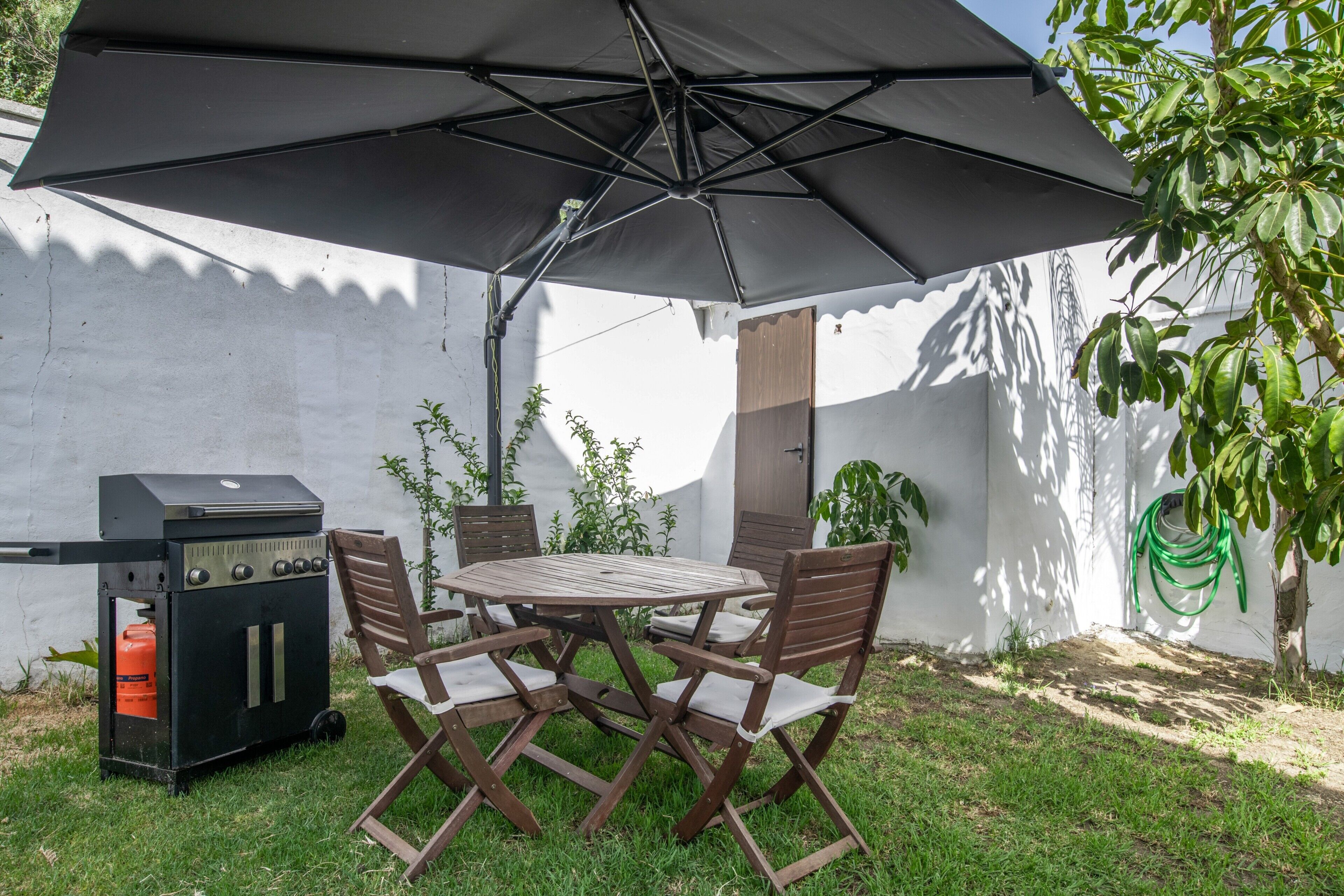 Um pátio externo com mesa redonda, quatro cadeiras, churrasqueira e um grande guarda-sol.