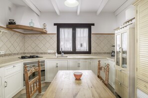 Een keuken met een houten tafel, witte kasten, een raam met gordijnen en een inbouwoven.