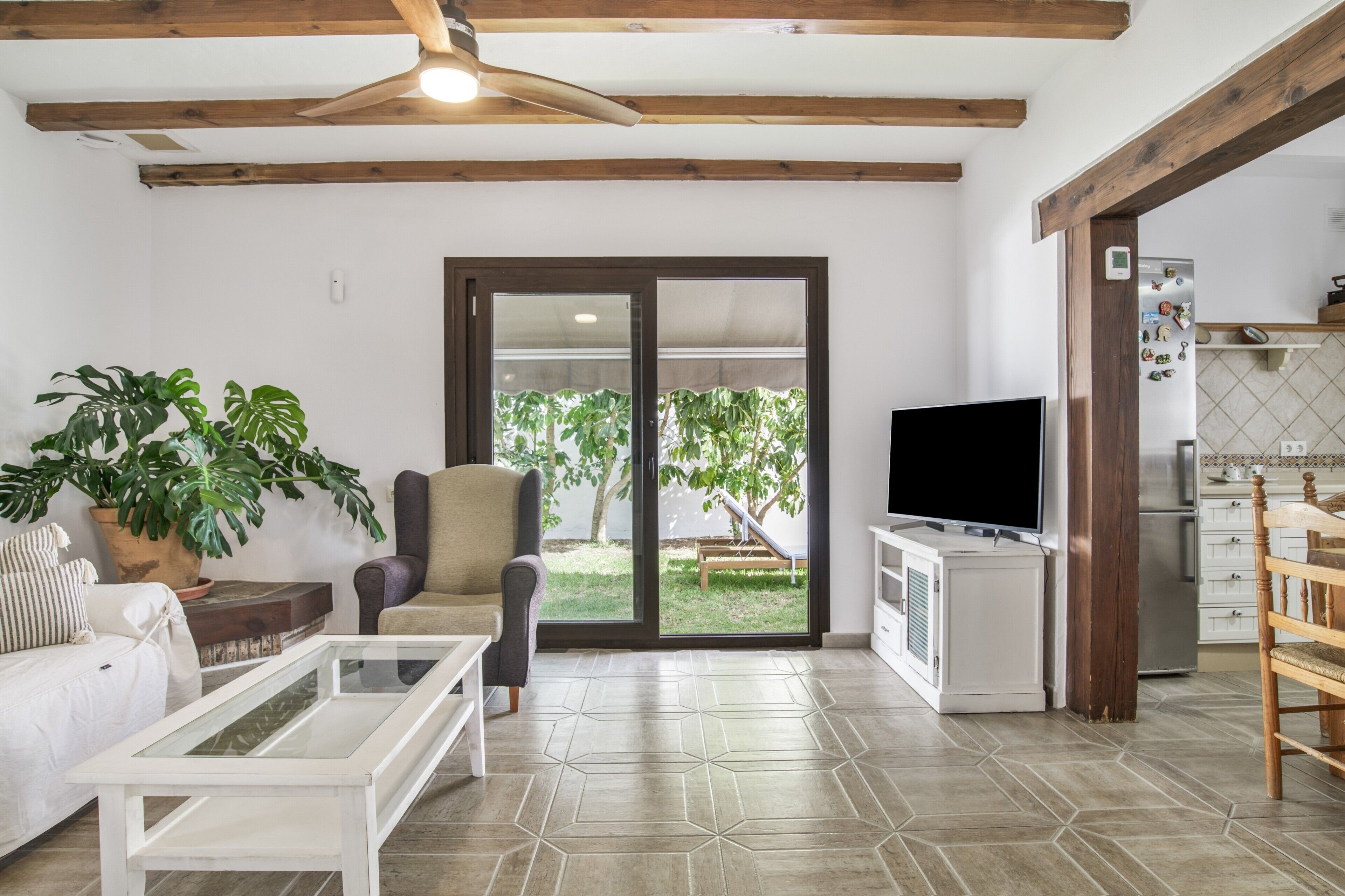 Ein Wohnzimmer mit Sofa, Sessel, Couchtisch, Fernseher und einer Küchenzeile mit Kühlschrank.
