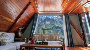 A cozy room with a wooden ceiling, a sofa, a table with decorative items, and a large window offering a mountain view. - Tiger Leaping Gorge yachajiao Manor (Lijiang)