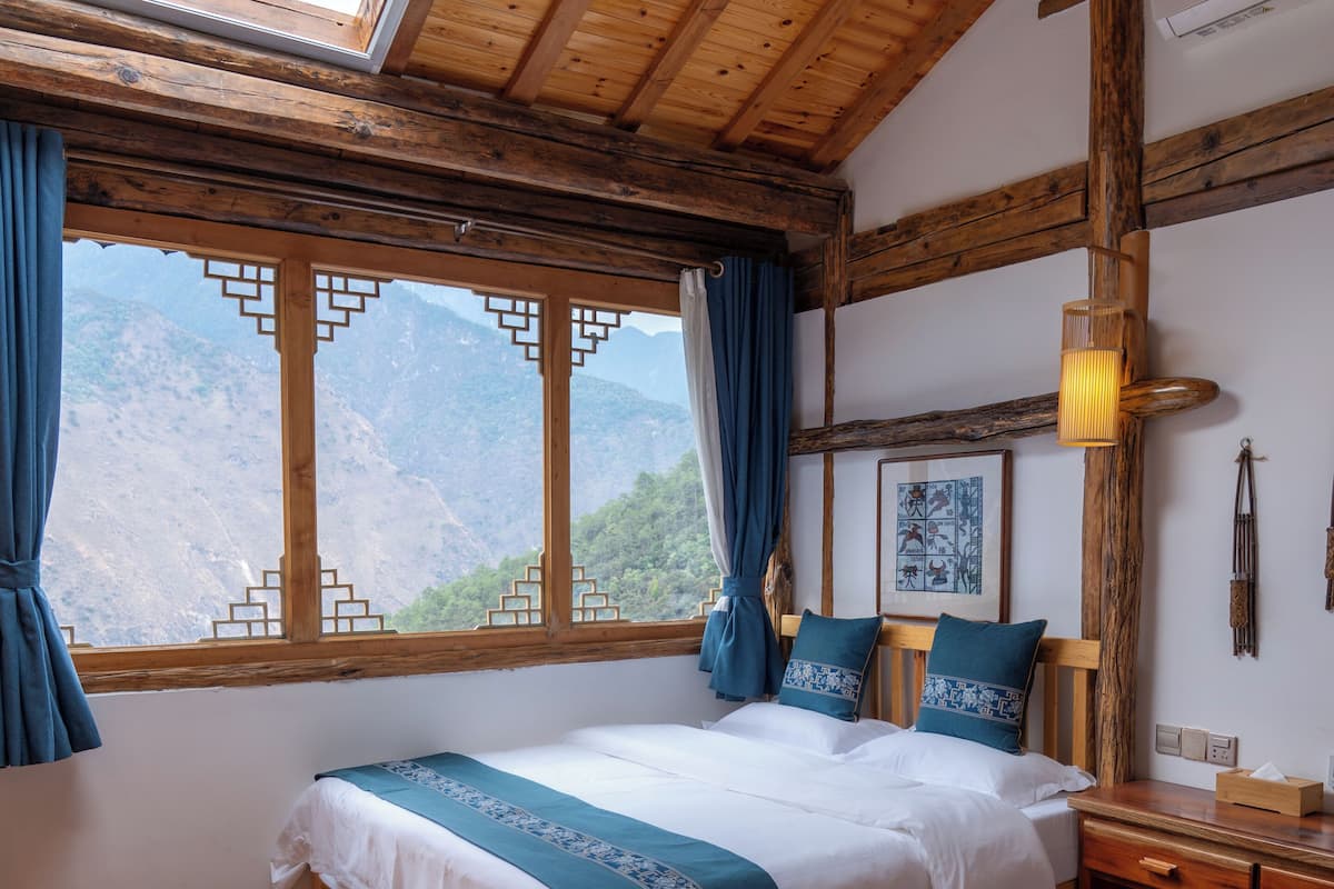 A room with a wooden ceiling, a bed with a blue and white bedspread, a nightstand, and a window with a view of mountains.